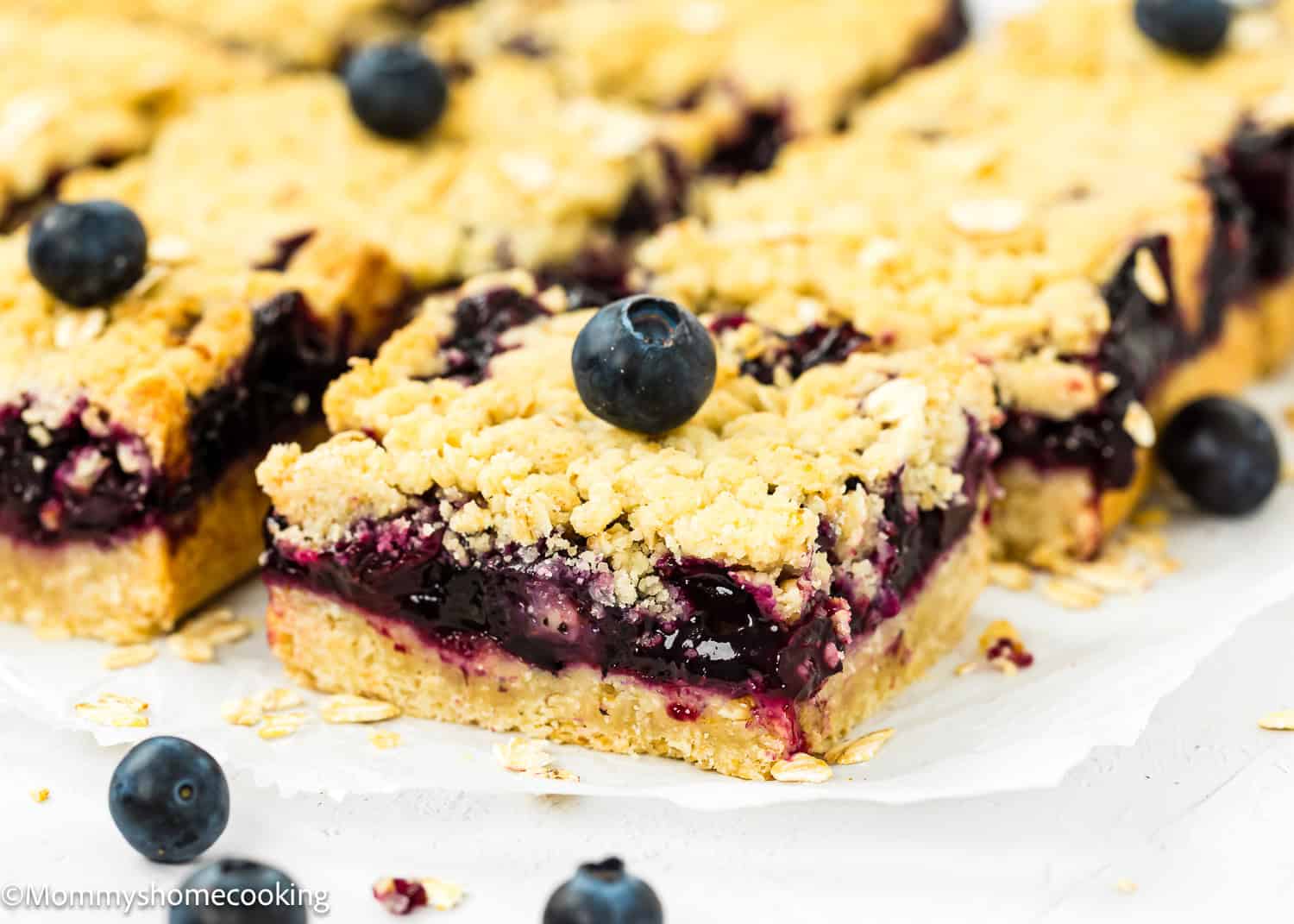 Easy eggless blueberry oat crumb bars on parchment paper, adorned with whole blueberries. The bars feature a crumbly top layer and visible blueberry filling.