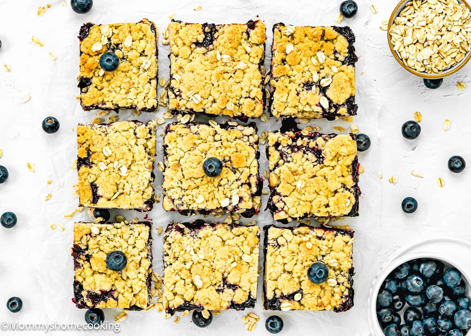 Nine easy eggless blueberry oat crumb bars topped with oats and a whole blueberry on each piece, arranged in a grid. Blueberries and oats are scattered around on a white surface.
