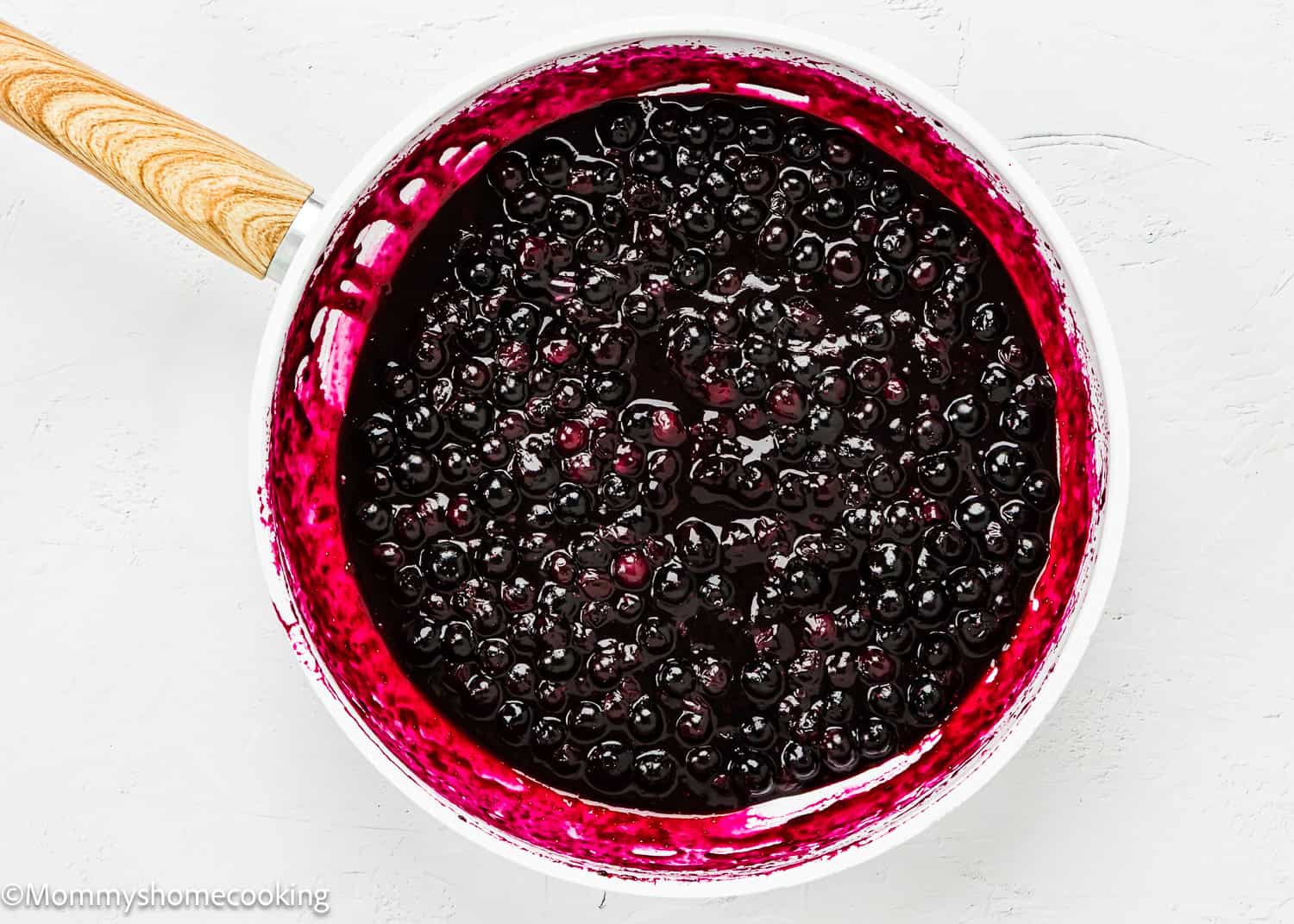 A white saucepan with a wooden handle contains a thick blue-purple sauce ready for Easy Eggless Blueberry Oat Crumb Bars.