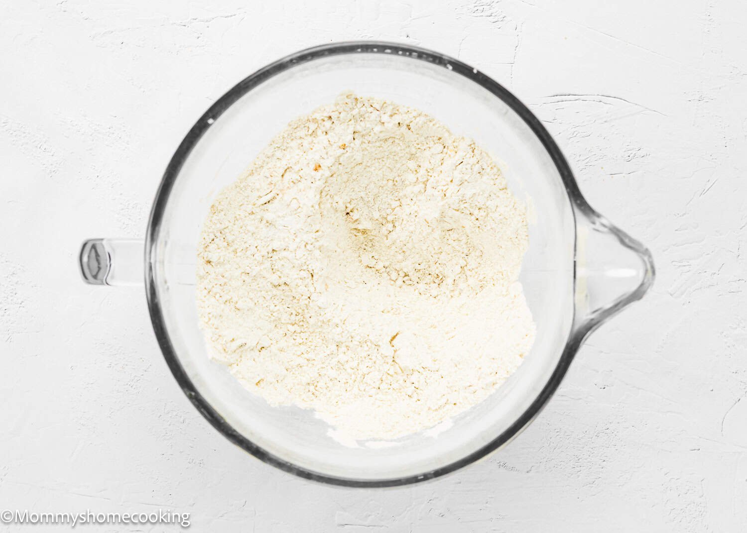 A glass bowl filled with a mound of sifted white flour sits on a white surface, ready to be transformed into easy eggless blueberry oat crumb bars.