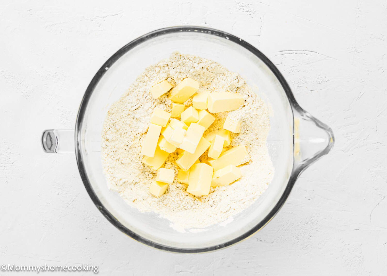A glass bowl containing flour, several cubes of butter, and ingredients to make easy eggless blueberry oat crumb bars sits on a white surface.