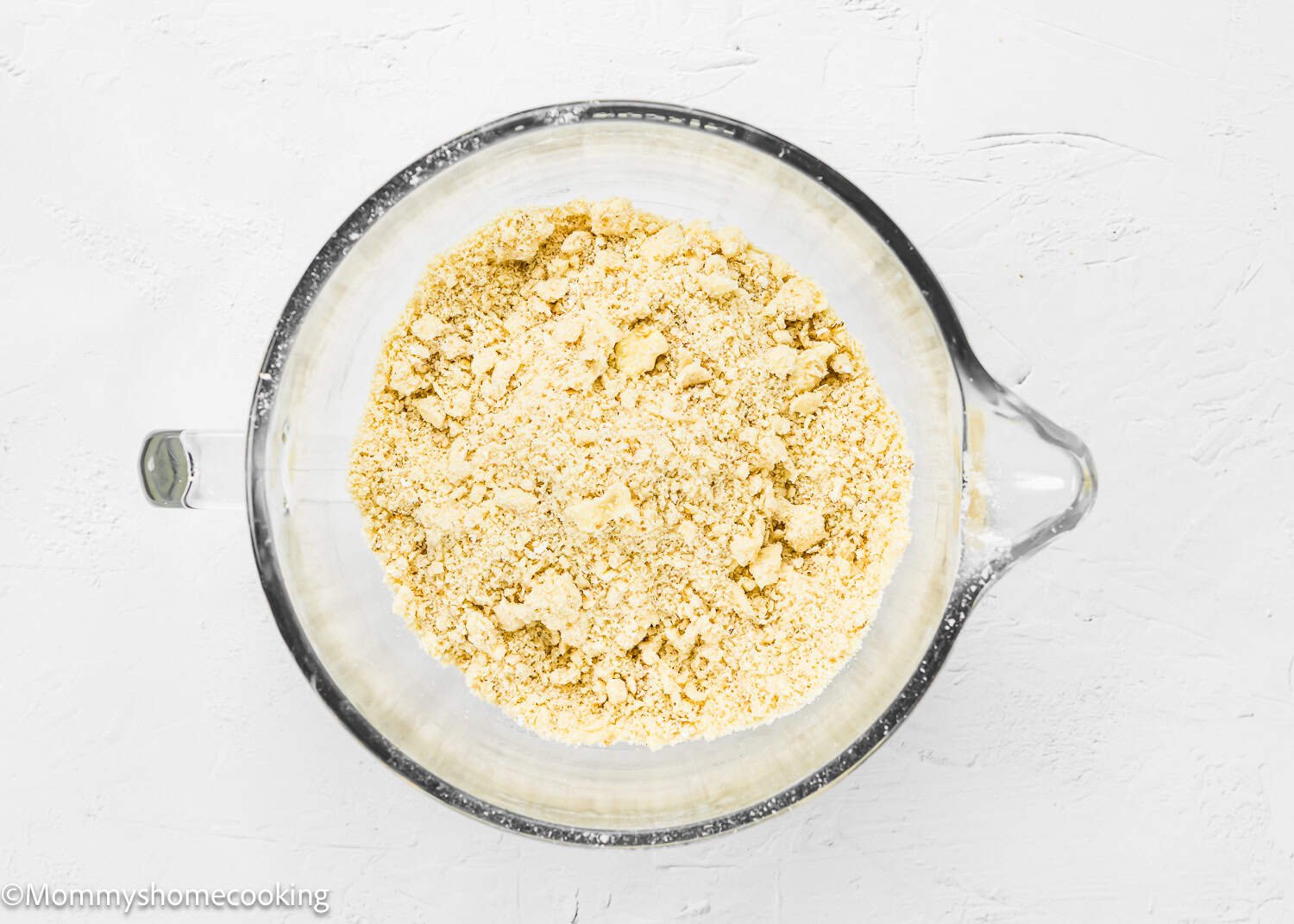 A glass bowl brimming with a crumbly yellow flour mixture sits on a white textured surface, ready to become the base for easy eggless blueberry oat crumb bars.