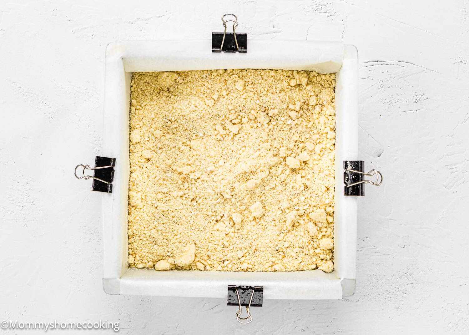 The square baking dish, secured with binder clips and lined with parchment paper, is filled with a crumbly yellow mixture on a textured white surface, ready to become delicious Easy Eggless Blueberry Oat Crumb Bars.