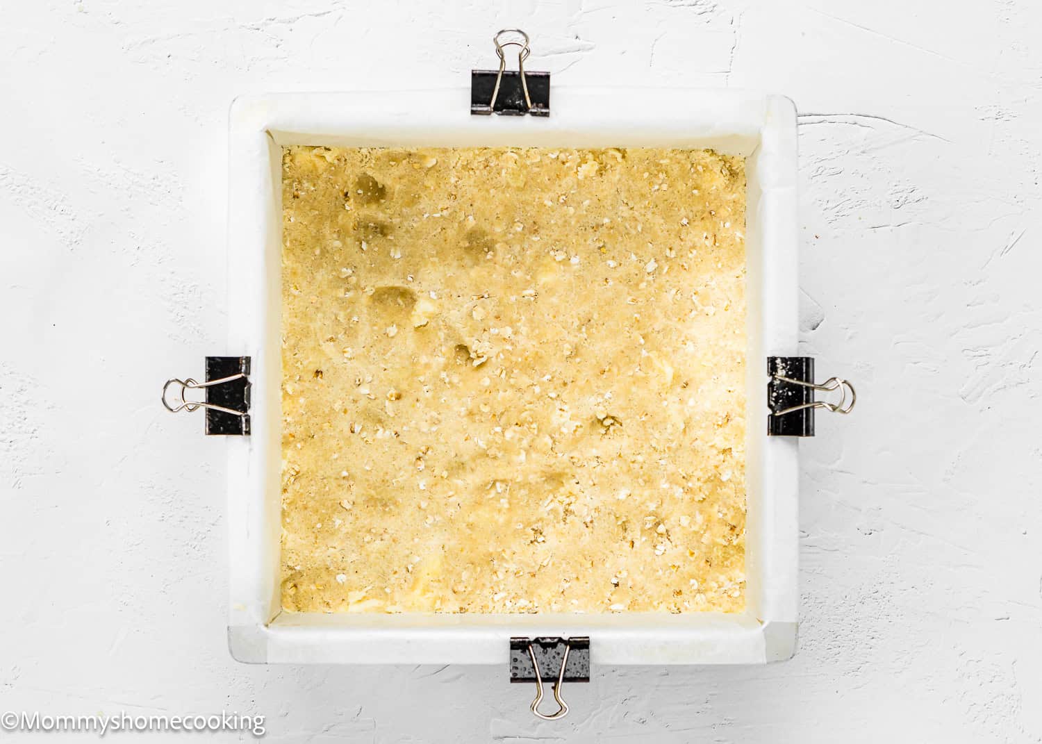 A square baking dish, lined with parchment paper and secured with binder clips, is filled with an unbaked crust mixture on a white textured surface. Perfect for making Easy Eggless Blueberry Oat Crumb Bars.