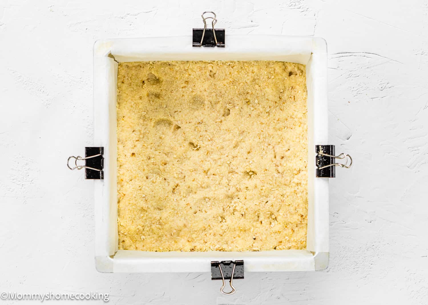 A square baking pan lined with parchment paper and held by clips, filled with an easy eggless blueberry oat crumbly dough mixture on a white surface.