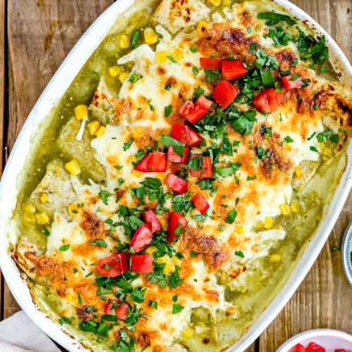 This easy baked casserole is topped with melted cheese, chopped tomatoes, and herbs. Corn kernels add a pop of color to the dish. Nearby, a small bowl of green sauce and half an avocado complete this delightful take on Easy Green Chicken Enchiladas on a wooden surface.