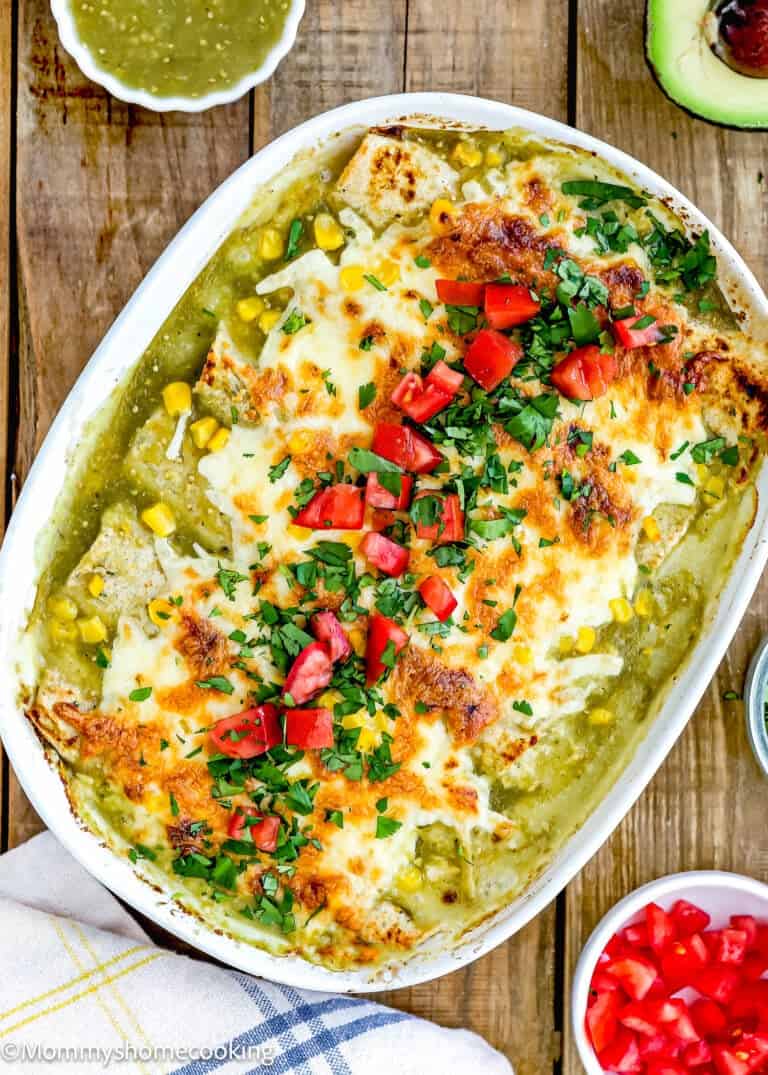 This easy baked casserole is topped with melted cheese, chopped tomatoes, and herbs. Corn kernels add a pop of color to the dish. Nearby, a small bowl of green sauce and half an avocado complete this delightful take on Easy Green Chicken Enchiladas on a wooden surface.
