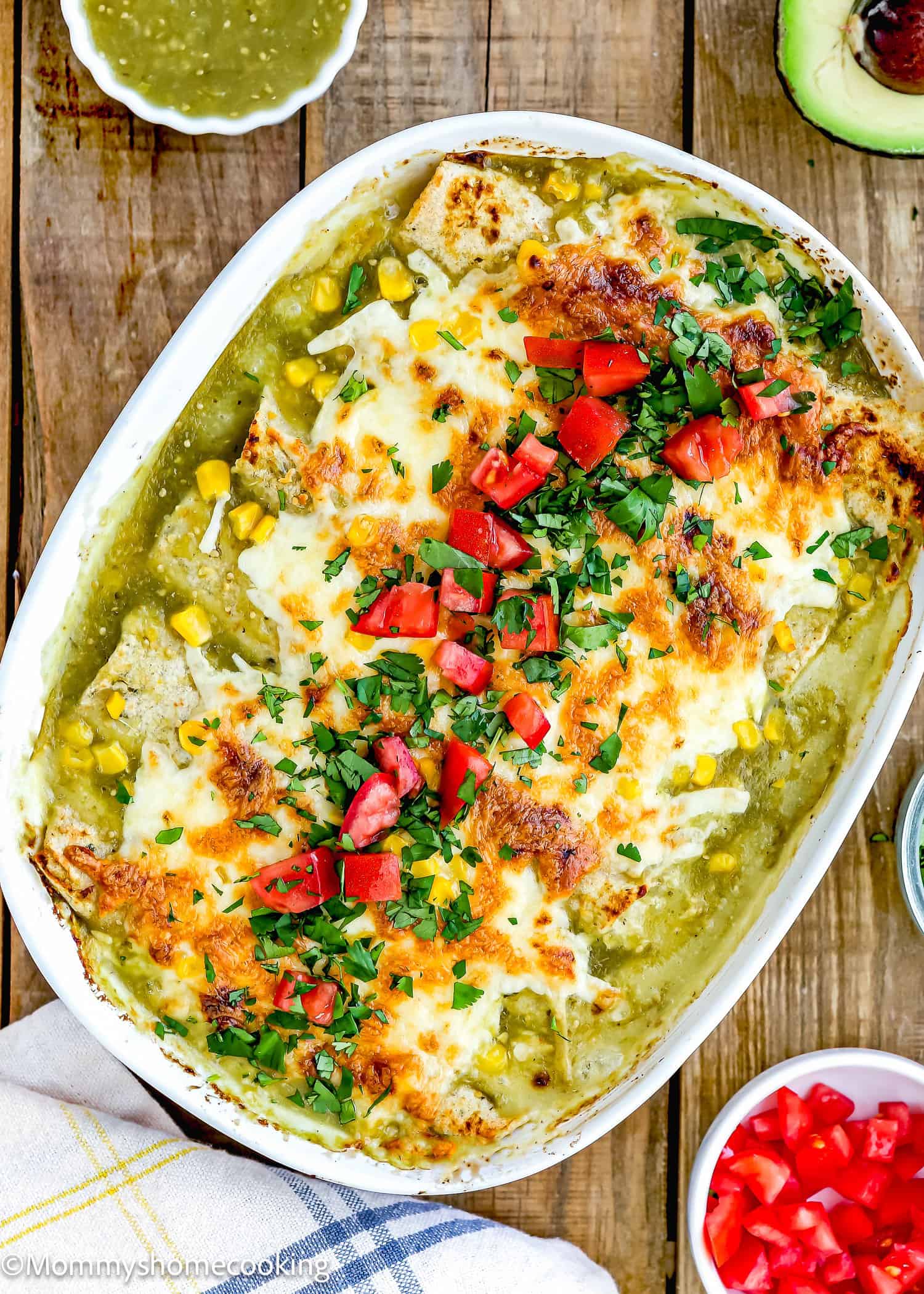 This easy baked casserole is topped with melted cheese, chopped tomatoes, and herbs. Corn kernels add a pop of color to the dish. Nearby, a small bowl of green sauce and half an avocado complete this delightful take on Easy Green Chicken Enchiladas on a wooden surface.