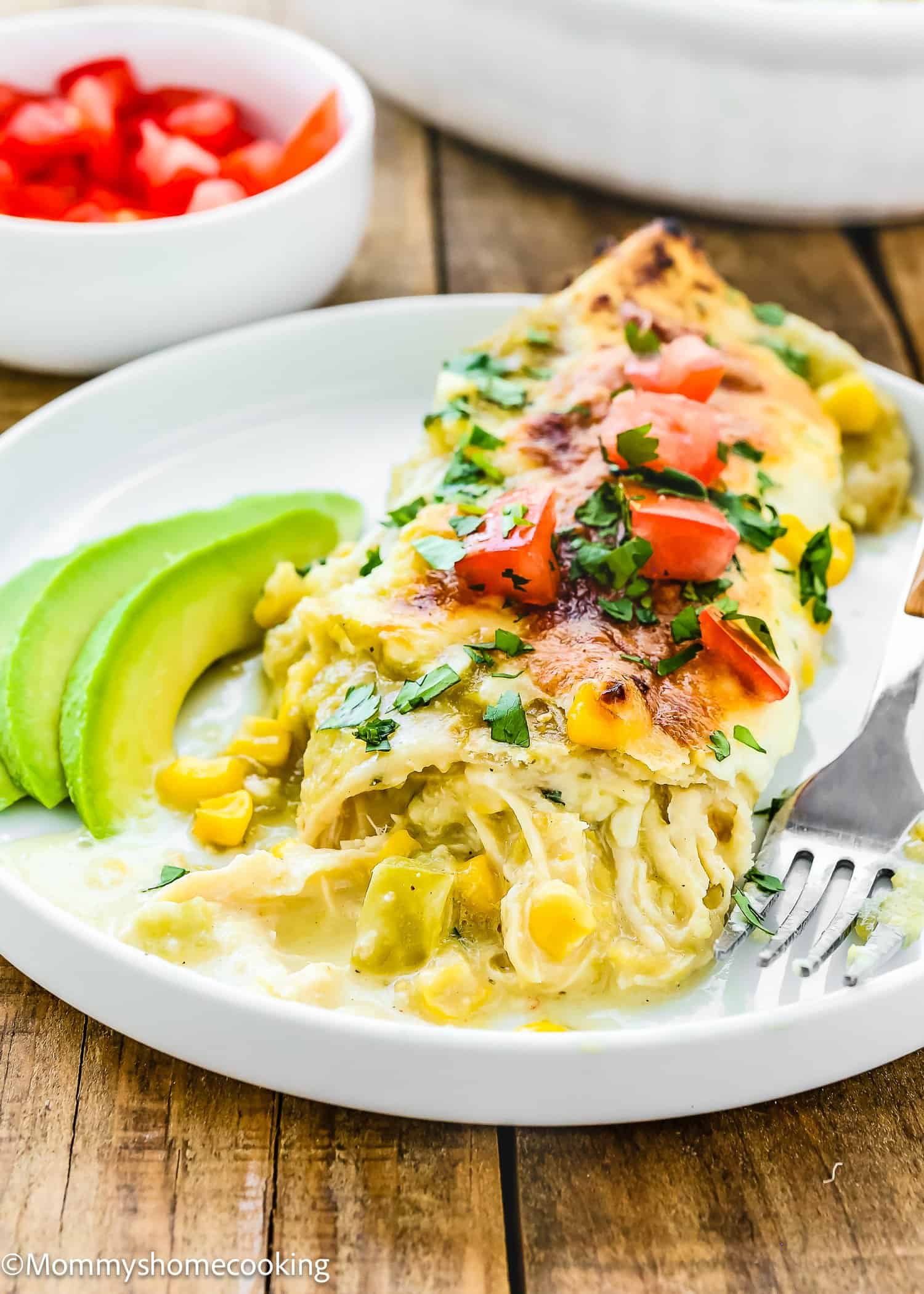 Enjoy an Easy Green Chicken Enchilada topped with chopped tomatoes and cilantro, accompanied by creamy avocado slices. A fork is on the side, ready to dig in, while a small bowl of diced tomatoes sits in the background.