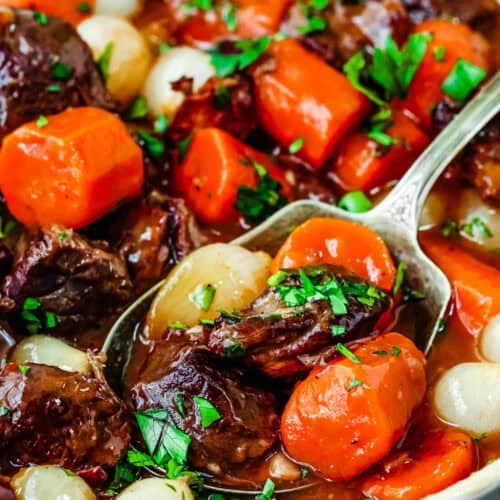 A close-up of Easy Instant Pot Beef Bourguignon with carrots, pearl onions, and herbs in a bowl. A spoon is scooping a portion.