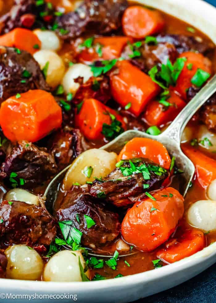 A close-up of Easy Instant Pot Beef Bourguignon with carrots, pearl onions, and herbs in a bowl. A spoon is scooping a portion.