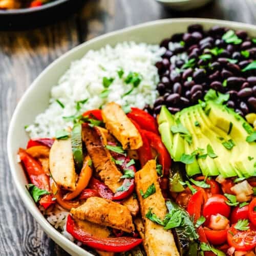 An easy turkey fajita bowl featuring grilled chicken strips, a medley of vibrant red and green peppers, creamy avocado, black beans, and rice topped with chopped tomatoes and fresh cilantro. This colorful dish is beautifully presented on a wooden table.