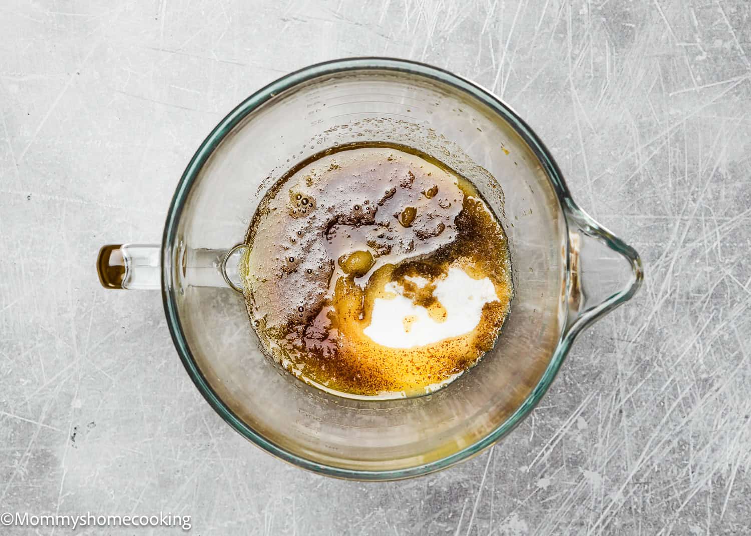 A glass measuring cup holds a mix of melted butter, brown sugar, and granulated sugar on a scratched gray surface – the perfect start for eggless brown butter snickerdoodle cookies.