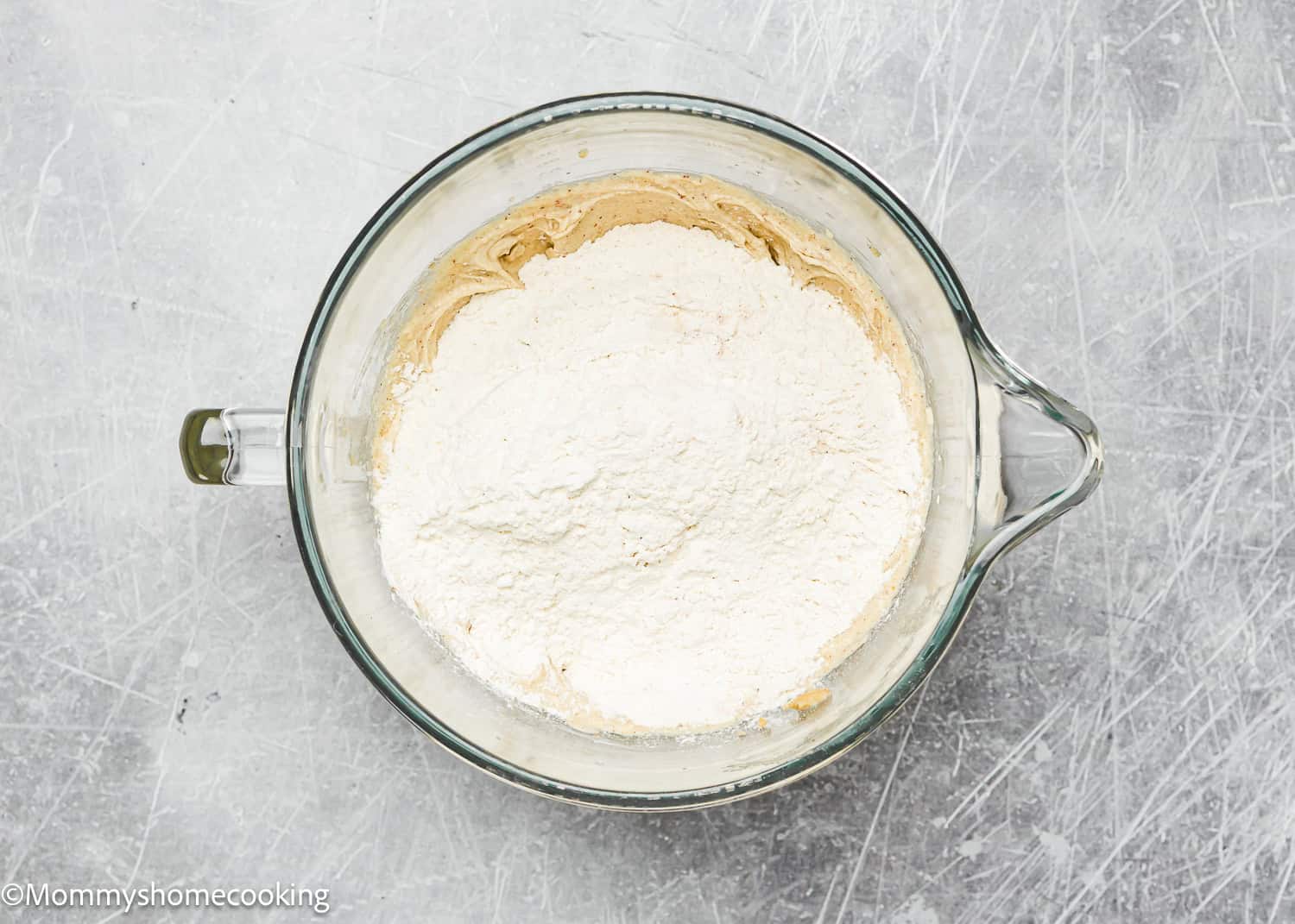 A glass mixing bowl containing dough for Eggless Brown Butter Snickerdoodle Cookies, with flour dusted on top, rests on a scratched metal surface.