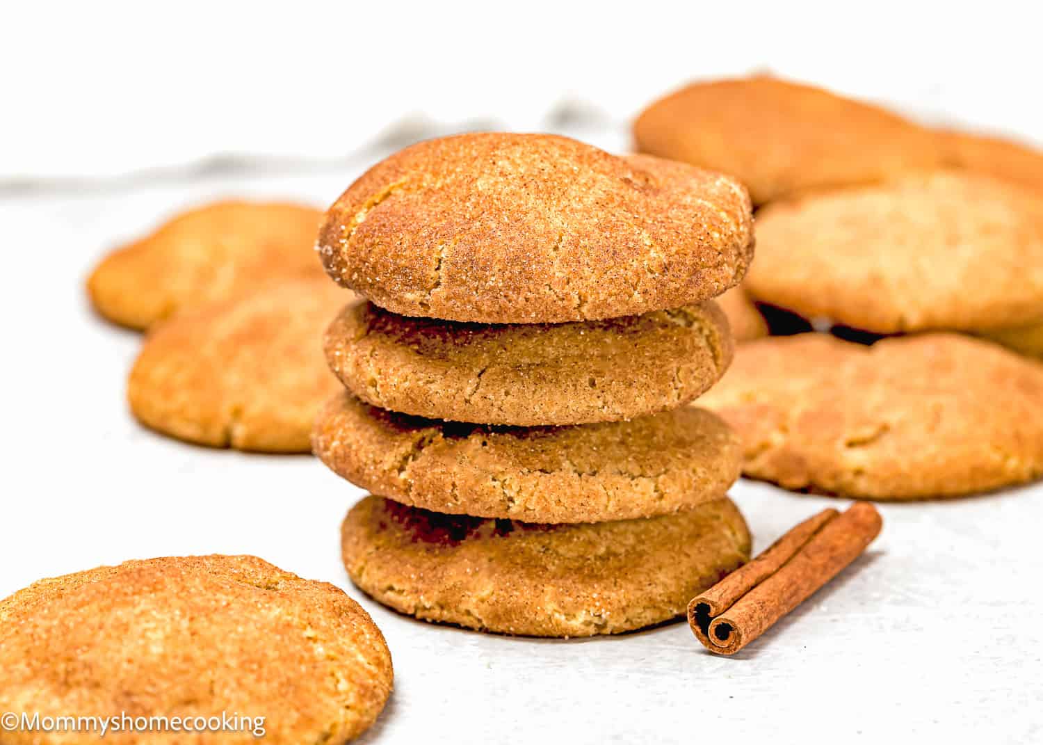 A stack of brown cookies, including Eggless Brown Butter Snickerdoodle Cookies, is surrounded by more cookies and a cinnamon stick on a white background.