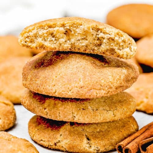 A stack of four eggless brown butter snickerdoodle cookies with the top one broken in half, revealing the interior. Two cinnamon sticks rest beside the stack, while more cookies appear in soft focus in the background.