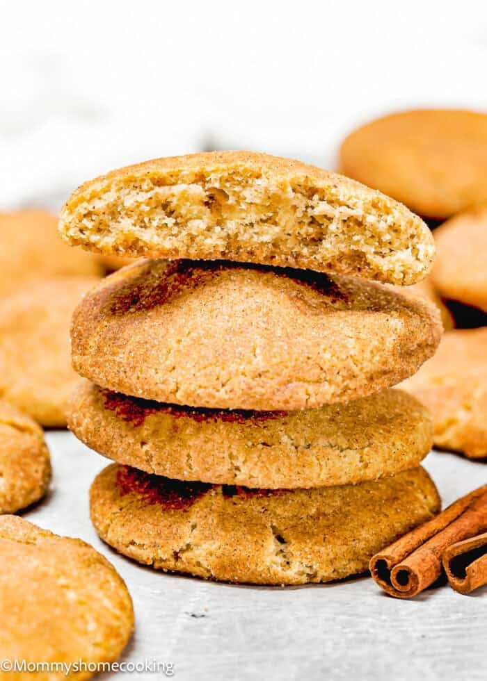 A stack of four eggless brown butter snickerdoodle cookies with the top one broken in half, revealing the interior. Two cinnamon sticks rest beside the stack, while more cookies appear in soft focus in the background.
