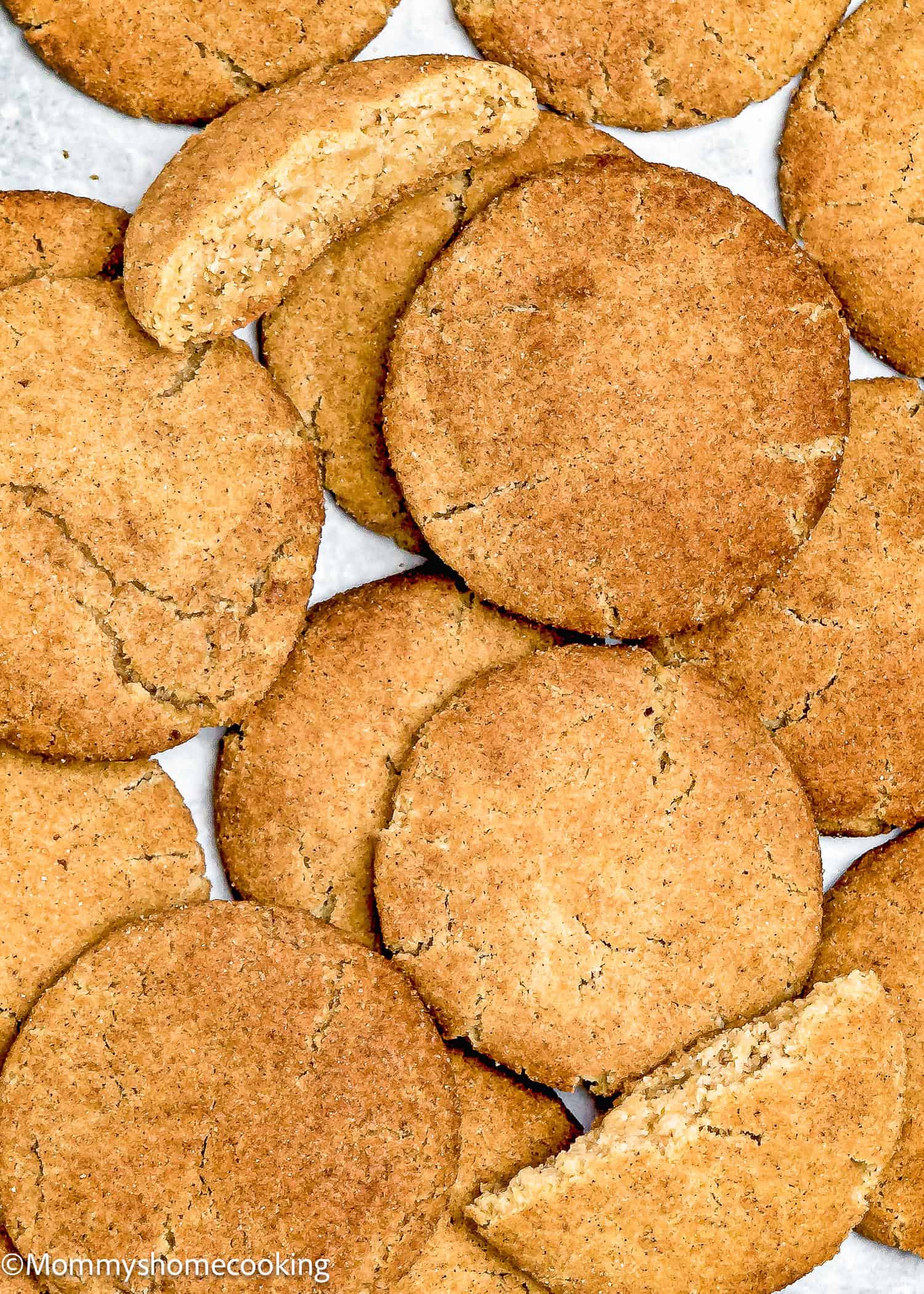 A pile of round, brown Eggless Brown Butter Snickerdoodle Cookies, some whole and some broken, sits on a white surface.