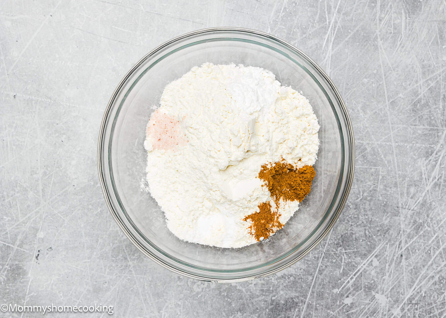 A glass bowl containing flour, baking powder, cinnamon, and a pinch of salt sits on a gray surface, ready for your eggless brown butter Snickerdoodle cookies.