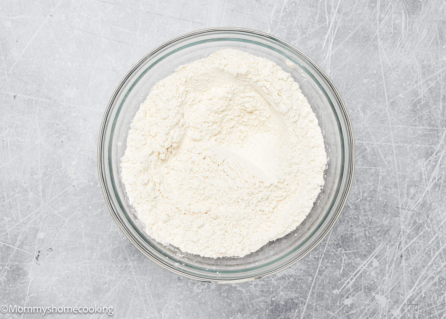 A clear glass bowl filled with white flour sits on a gray textured surface, ready for mixing ingredients to make eggless brown butter snickerdoodle cookies.