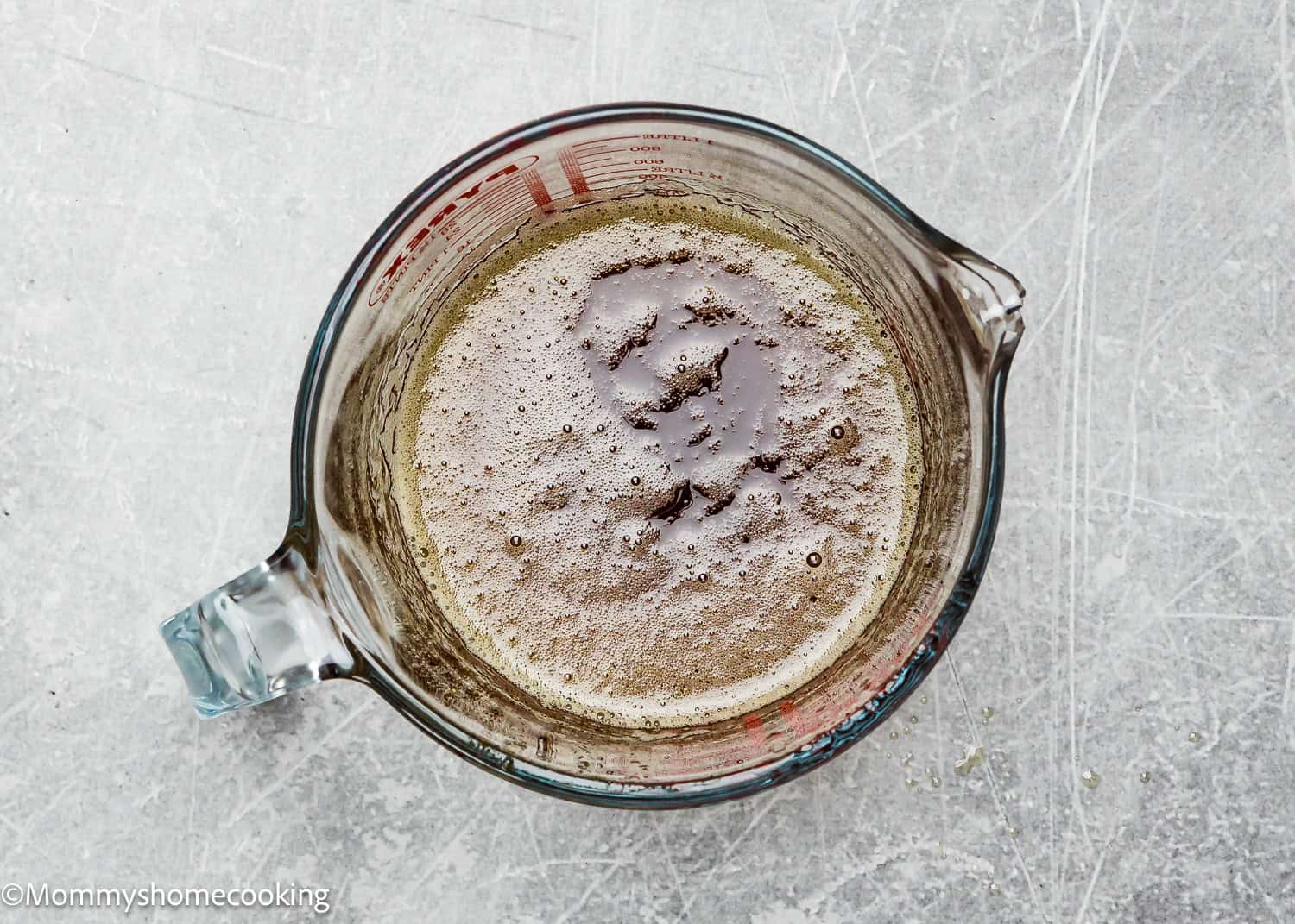 Overhead view of a glass measuring cup filled with brown butter on a gray textured surface, perfect for preparing eggless brown butter snickerdoodle cookies.