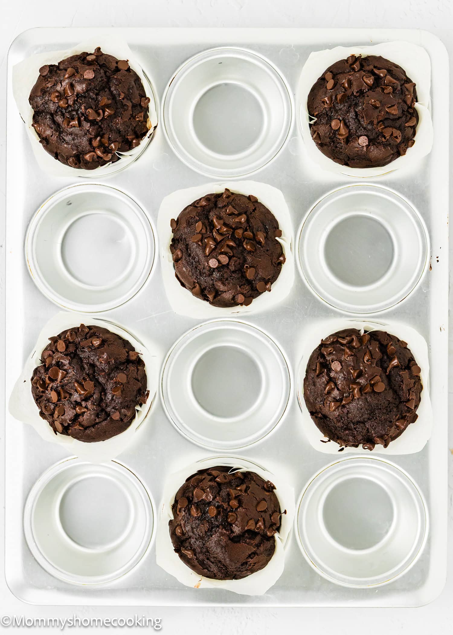 A muffin tray holds six baked eggless chocolate zucchini muffins topped with chocolate chips, placed in alternating slots. Three slots are empty.