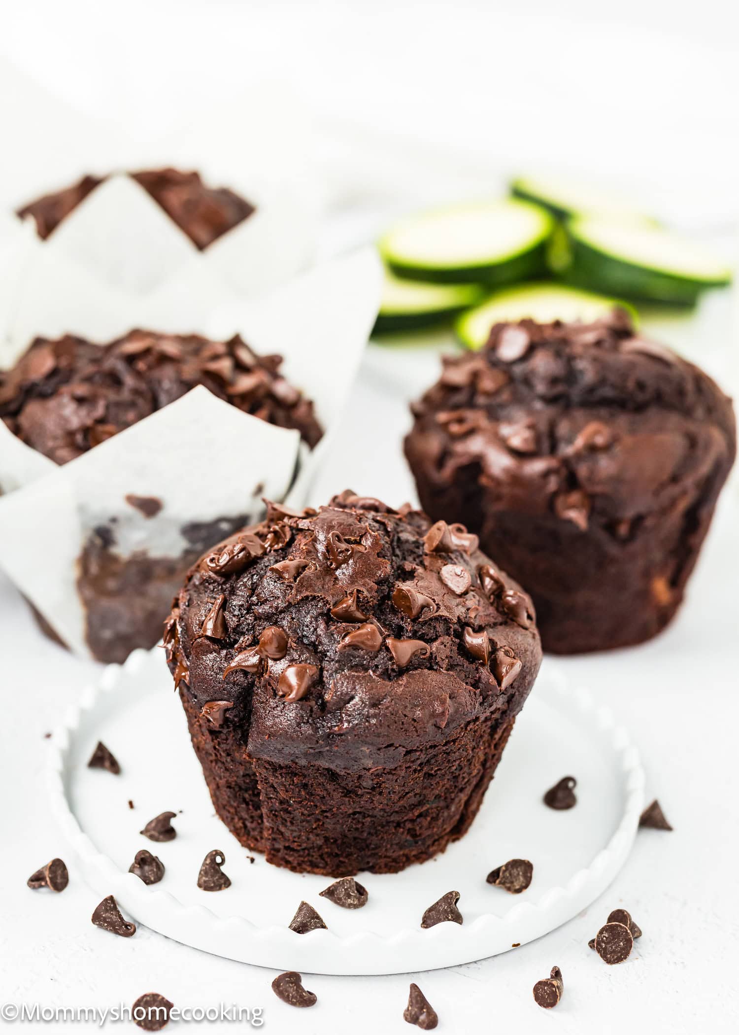 Eggless chocolate zucchini muffins topped with chocolate chips rest on a white plate. Sliced zucchini is blurred in the background.