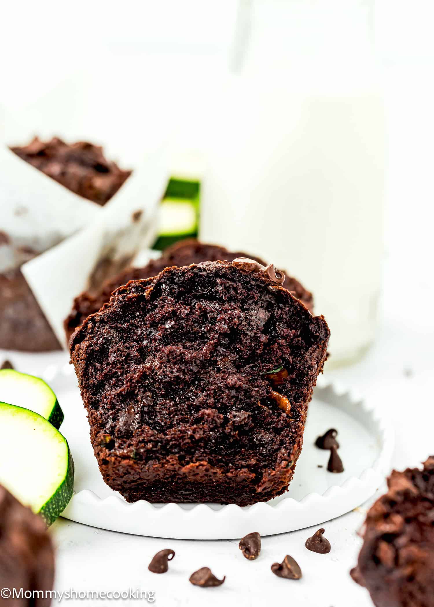 An eggless chocolate zucchini muffin, sliced on a paper liner, is surrounded by chocolate chips and zucchini slices, with a glass of milk in the background.