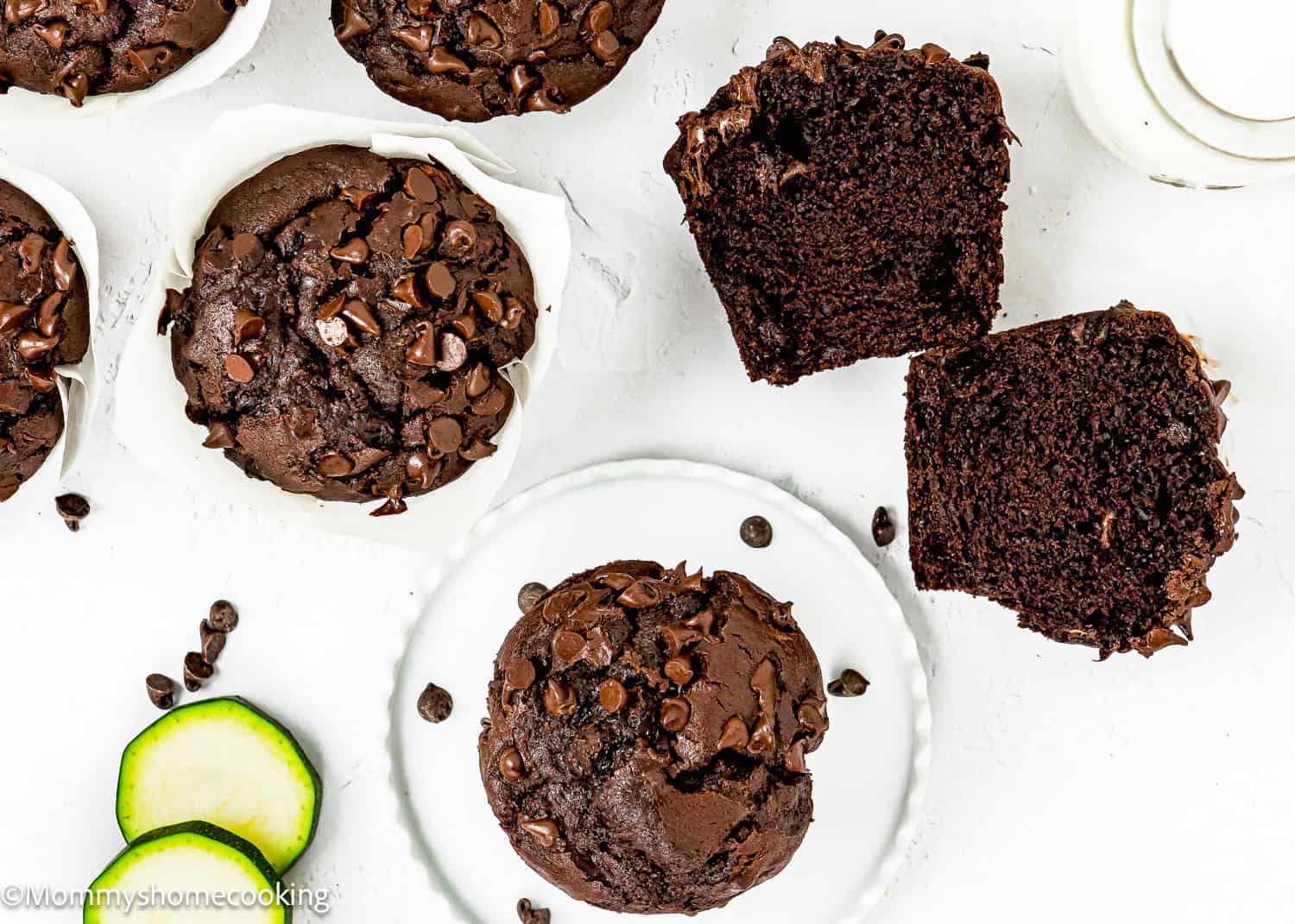 Eggless chocolate zucchini muffins topped with chocolate chips, one sliced in half, displayed on a white surface with fresh zucchini slices nearby.