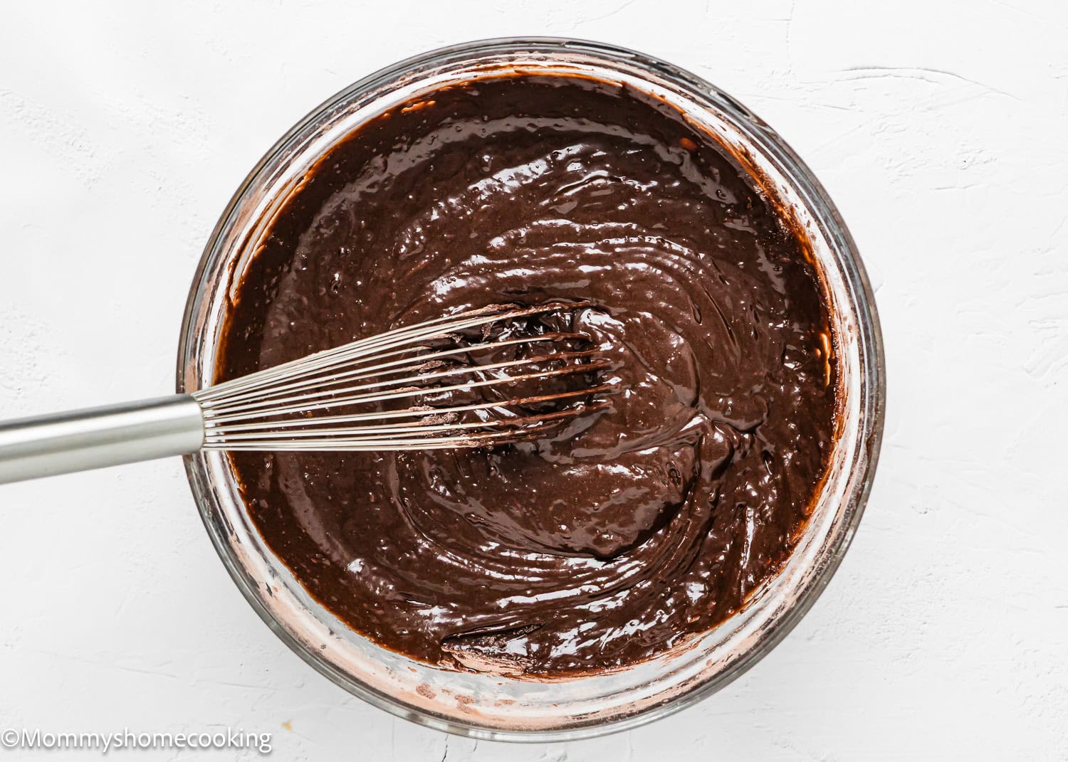 A metal whisk stirs a glass bowl of thick, dark chocolate batter on a white surface, ready to bake into delicious eggless chocolate zucchini muffins.