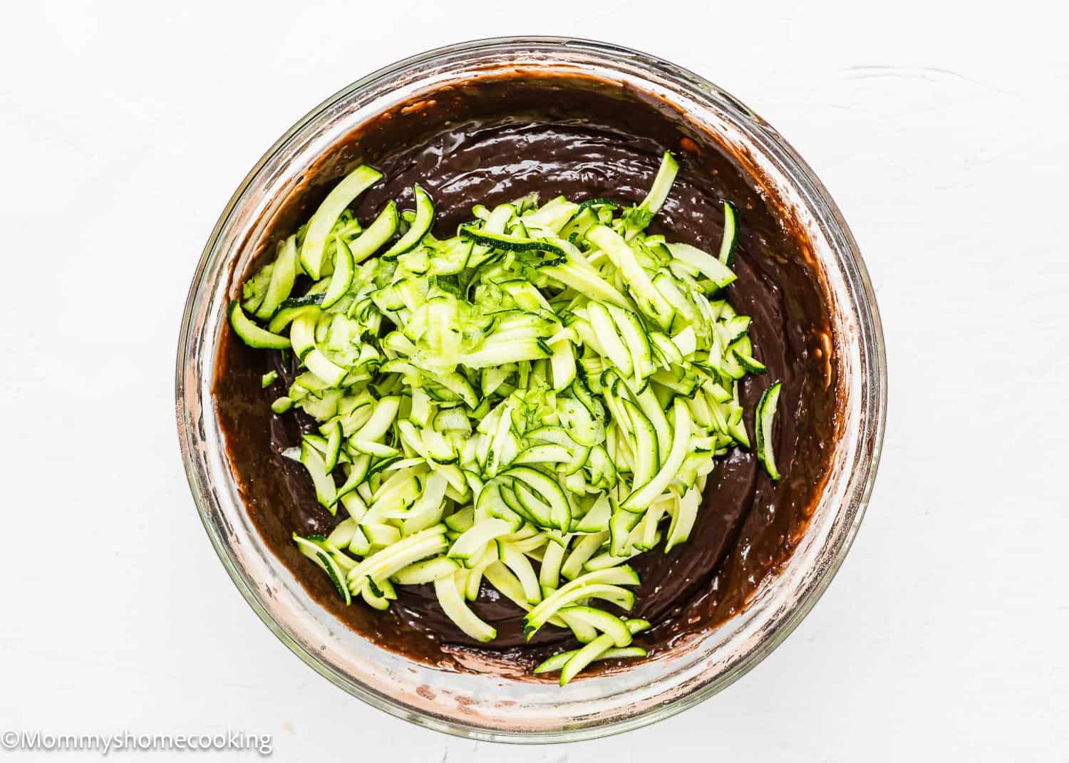 A bowl filled with rich chocolate batter topped with fresh sliced zucchini pieces on a light background, ready to transform into delectable eggless chocolate zucchini muffins.