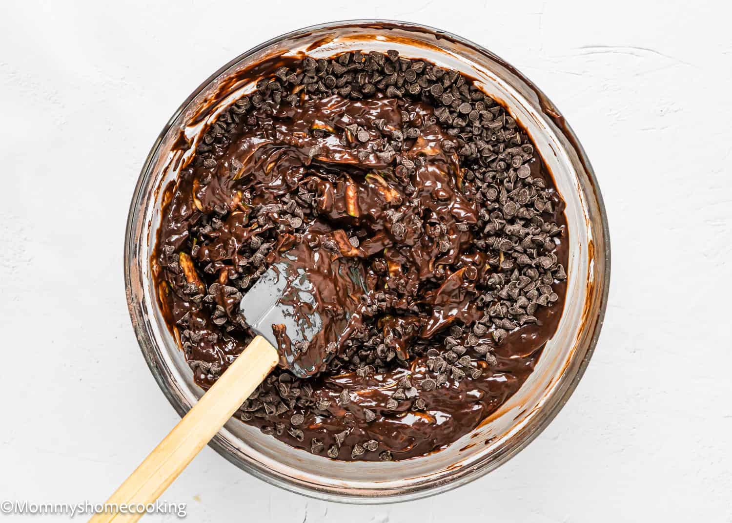A bowl of eggless zucchini chocolate batter mixture mixed with chocolate chips, being stirred with a spatula, perfect for making delicious zucchini muffins.