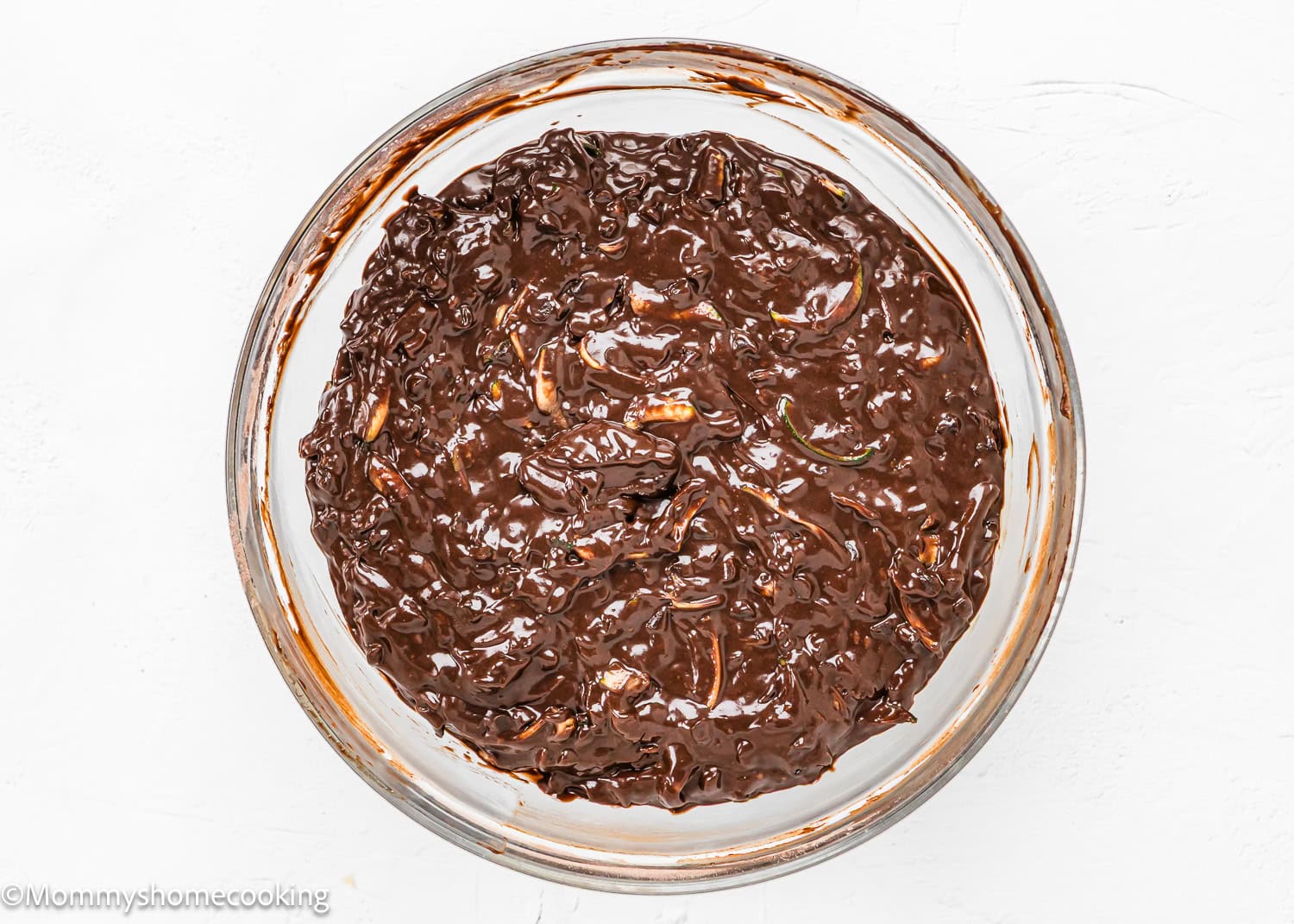 A glass bowl filled with eggless zucchini chocolate muffin batter, rests on a white surface.