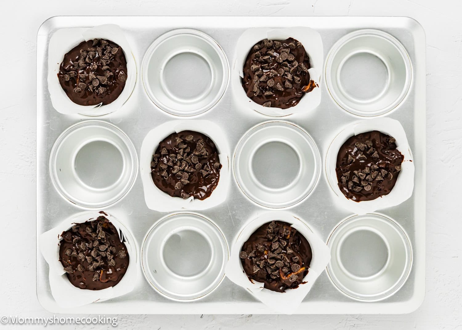 Muffin pan with six paper liners filled with rich chocolate batter and chocolate chips, offering a delightful preparation for Eggless Chocolate Zucchini Muffins. Three cups remain empty, hinting at more deliciousness to come.