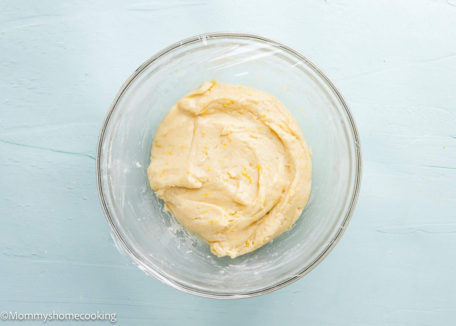 A bowl of creamy dough for Eggless Lemon Crinkle Cookies rests on a light blue surface, covered with plastic wrap.