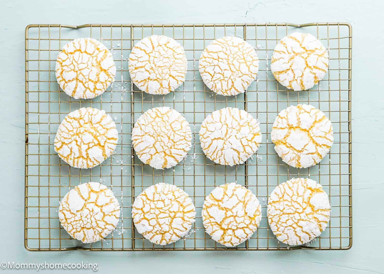 A dozen eggless lemon crinkle cookies with powdered sugar on top are arranged in three rows of four, cooling on a wire rack.