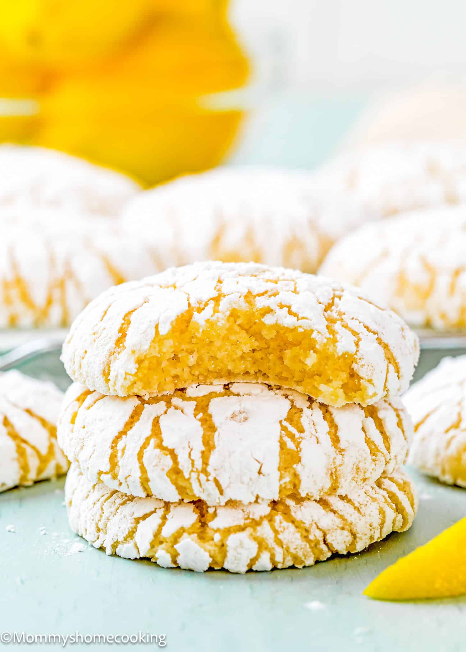 Three eggless lemon crinkle cookies stacked on a light green surface, with the top cookie partially bitten. More cookies and lemons are blurred in the background.