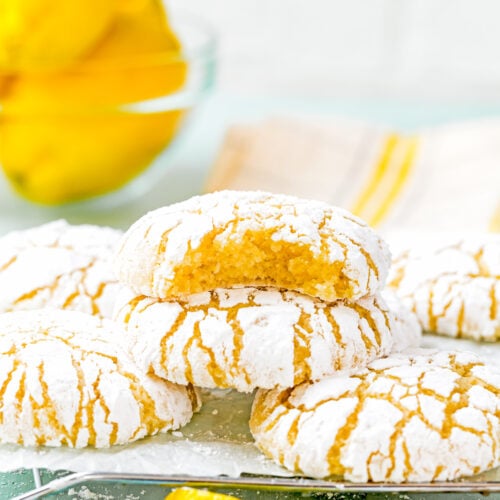A stack of eggless lemon crinkle cookies dusted with powdered sugar sits temptingly, one partially eaten, while fresh lemons add a burst of color in the background.
