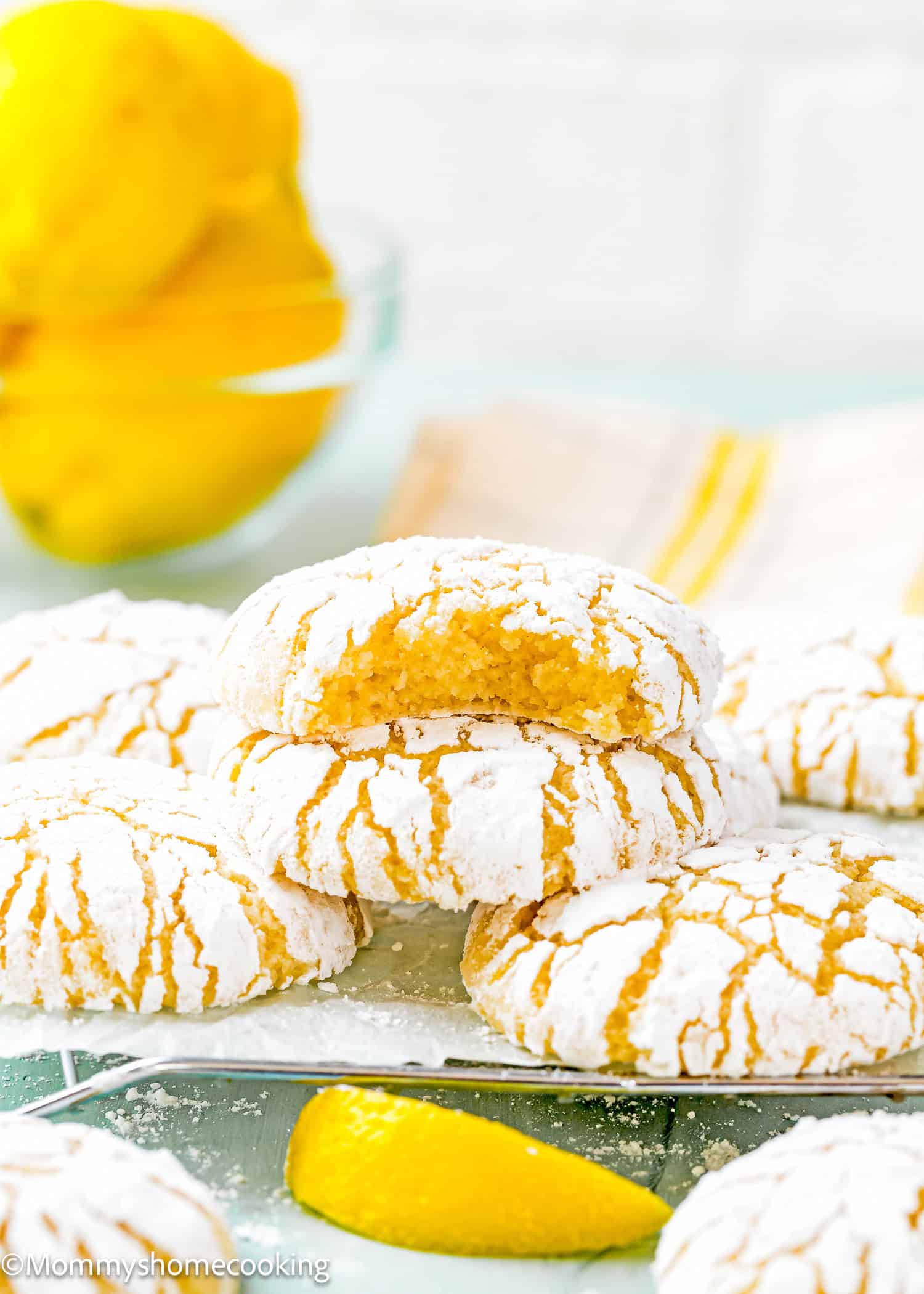 A stack of eggless lemon crinkle cookies dusted with powdered sugar sits temptingly, one partially eaten, while fresh lemons add a burst of color in the background.