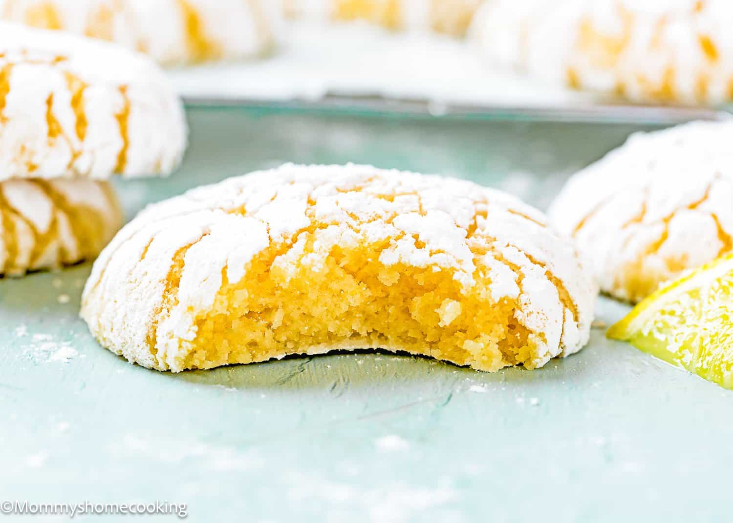 An eggless lemon crinkle cookie with a bite taken out, dusted with powdered sugar, sits surrounded by other cookies and a lemon slice on a vibrant green surface.