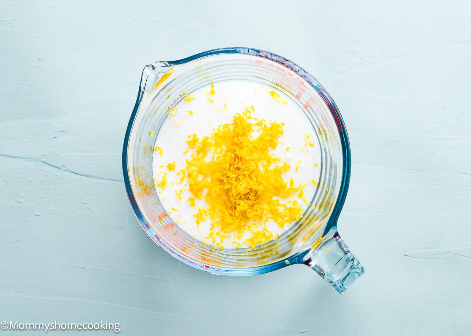 A glass measuring cup holds milk with a heap of yellow lemon zest on top, situated on a light blue surface, ideal for making Eggless Lemon Crinkle Cookies.