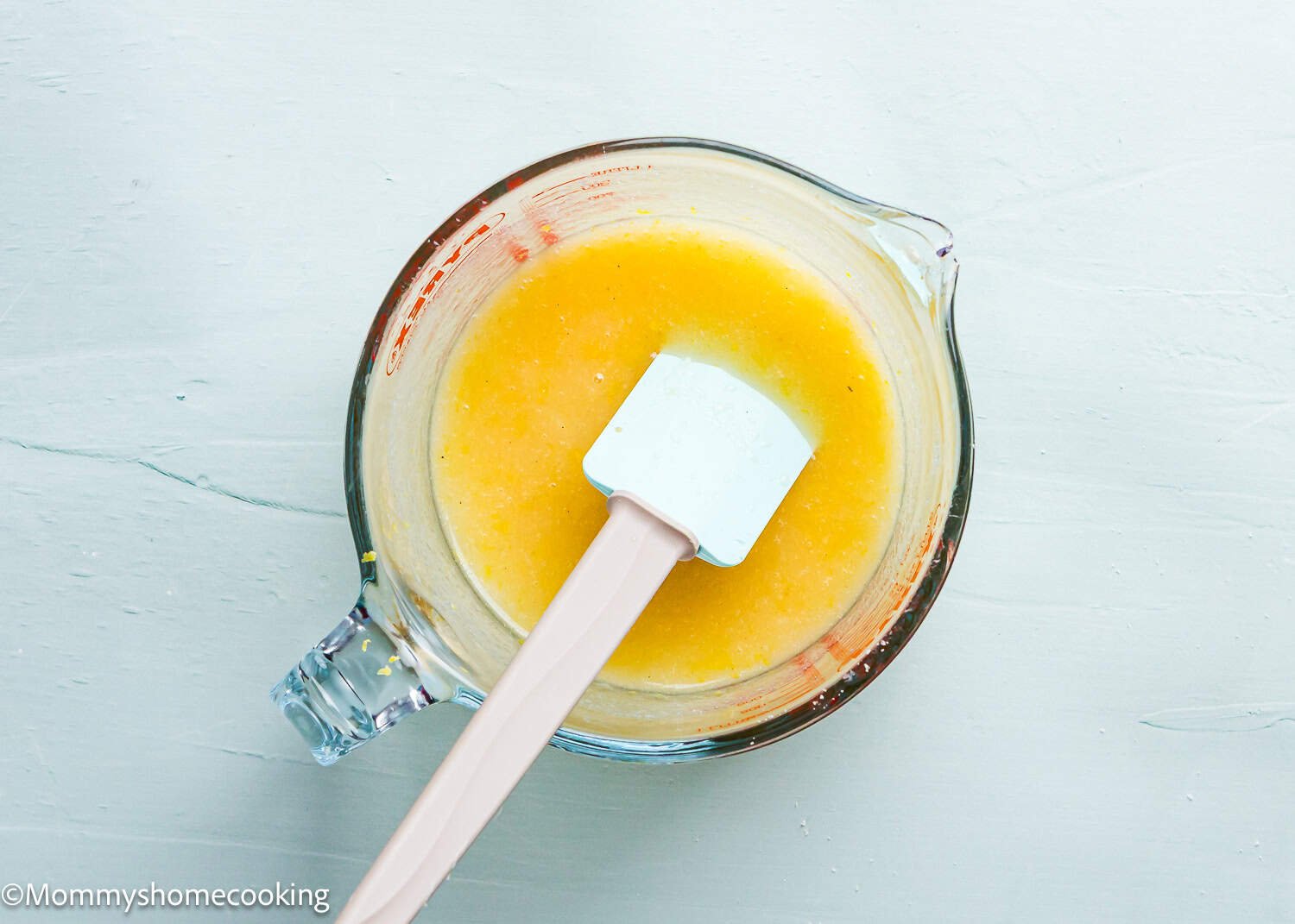 A glass measuring cup filled with a bright yellow mixture for Eggless Lemon Crinkle Cookies and a white spatula resting nearby on a light background.