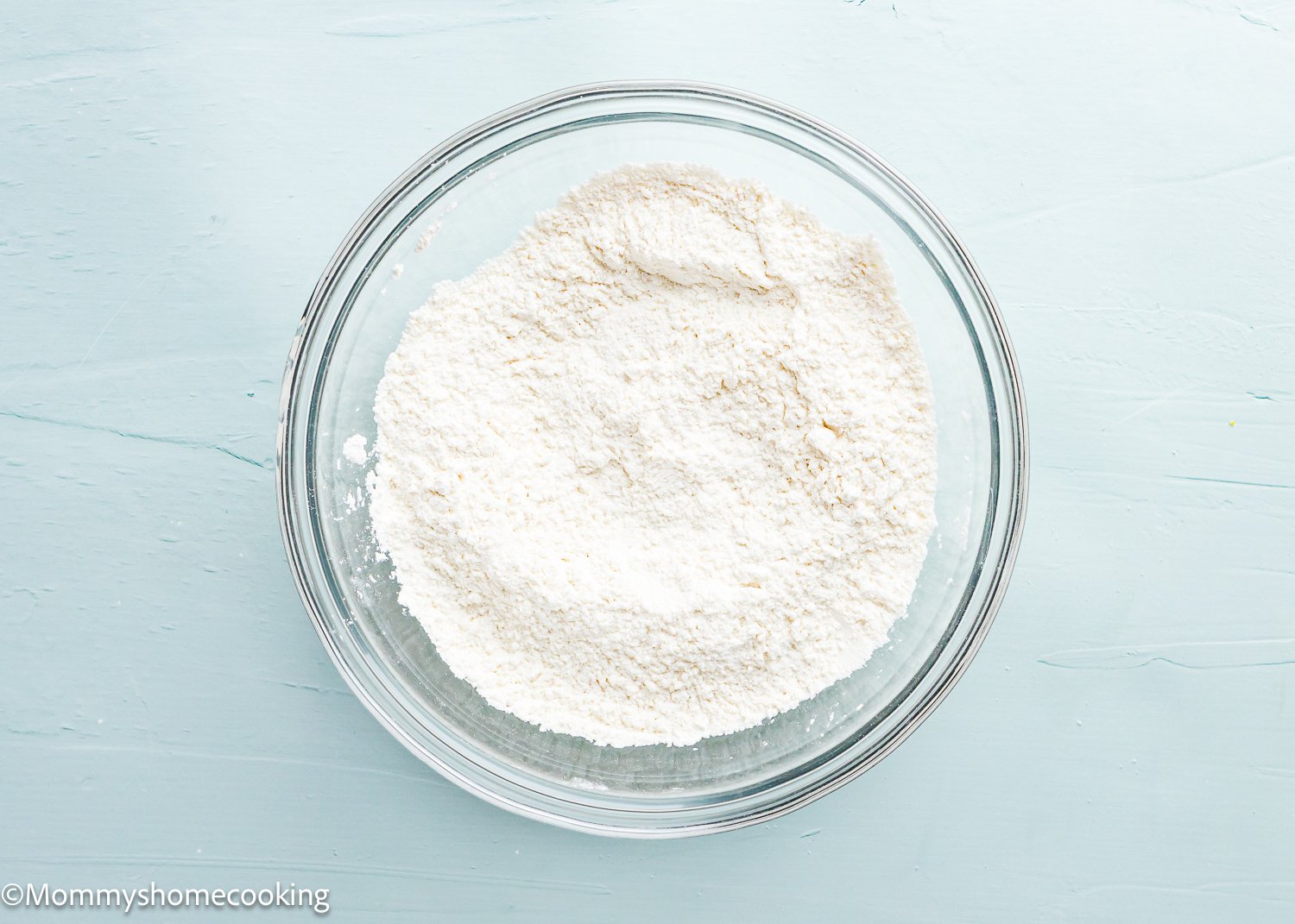 A glass bowl filled with a white powdery substance, perfect for making eggless lemon crinkle cookies, is placed on a light blue surface.