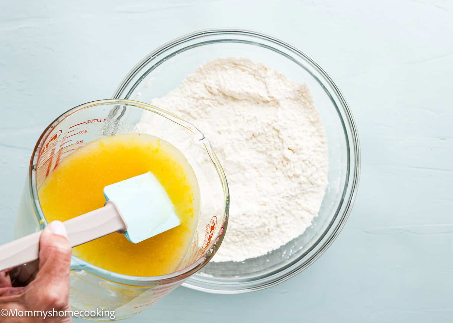 A person is making eggless lemon crinkle cookies by pouring yellow liquid into a bowl of flour with a glass measuring cup and spatula.