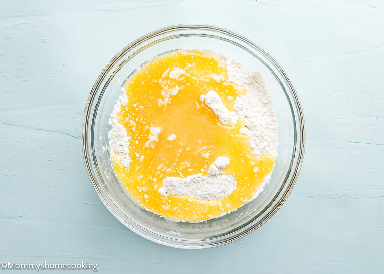 A clear glass bowl on a light blue surface contains ingredients for eggless lemon crinkle cookies: flour, melted butter, and milk.