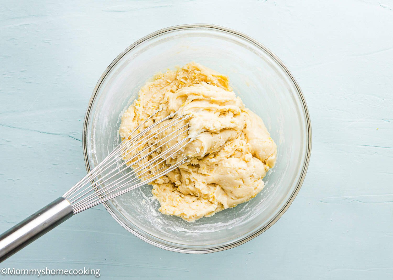 A whisk in a glass bowl with dough for eggless lemon crinkle cookies on a light blue surface.