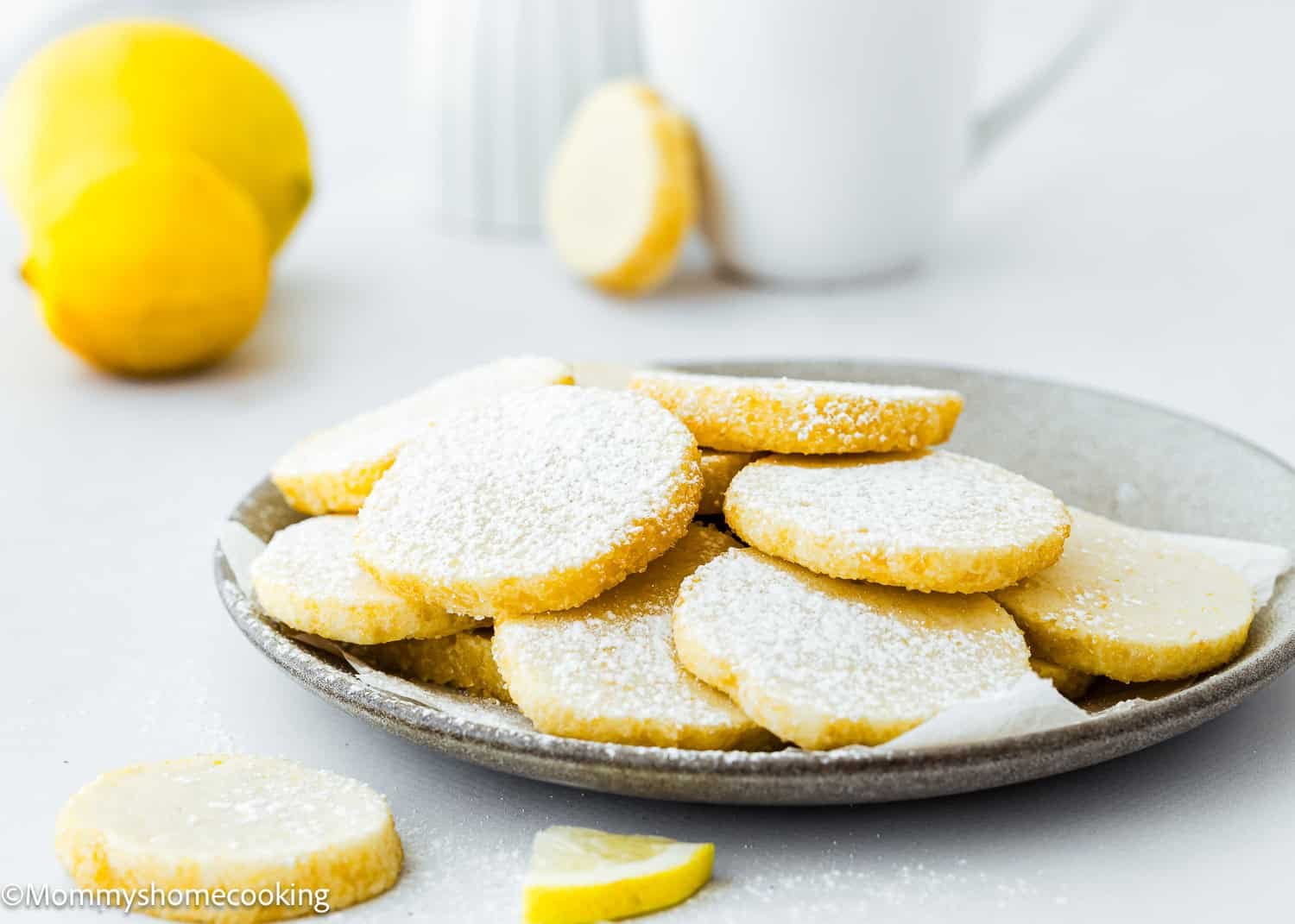 A plate of powdered sugar-dusted eggless lemon slice and bake cookies sits on the table, surrounded by fresh lemons and a pristine white mug in the background.