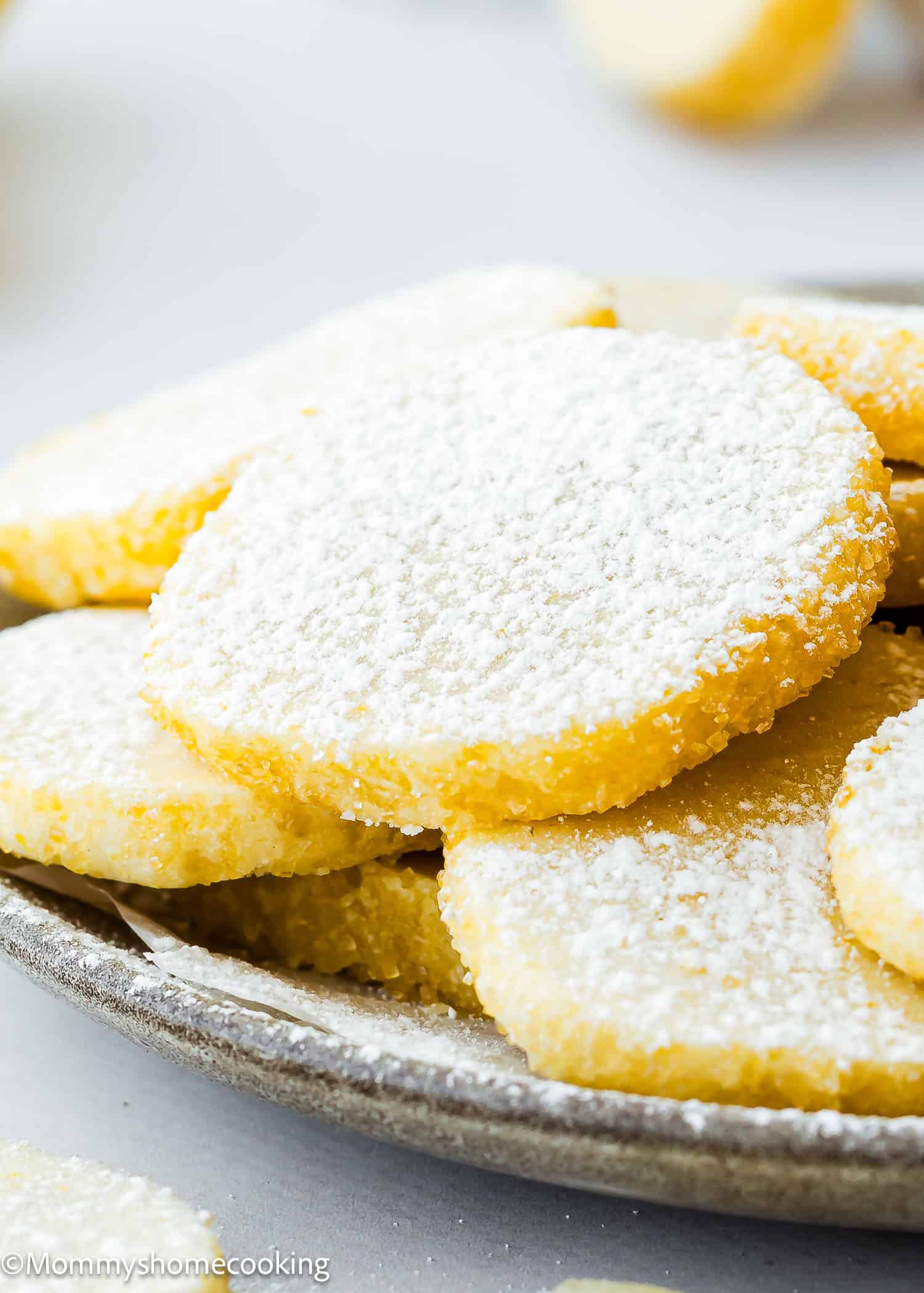 A plate of round, eggless lemon slice and bake cookies is artfully sprinkled with powdered sugar, offering a delightful citrus twist.
