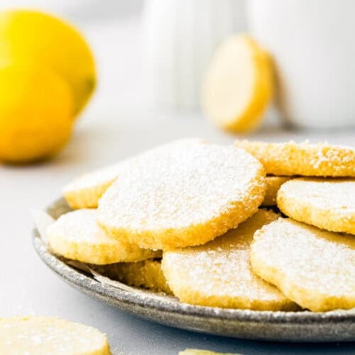 A plate of round, powdered sugar-coated cookies is on a table, embodying the delightful essence of eggless lemon slice and bake cookies. A fresh lemon and a cup are in the background, enhancing the scene with a touch of zest and warmth.