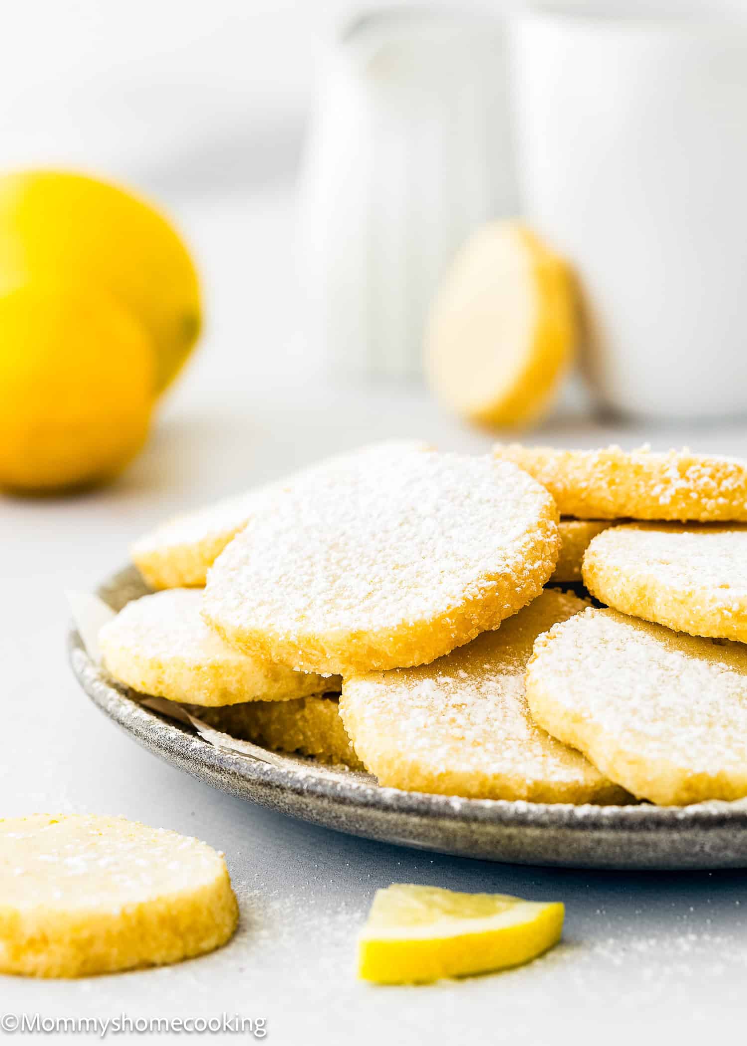 A plate of round, powdered sugar-coated cookies is on a table, embodying the delightful essence of eggless lemon slice and bake cookies. A fresh lemon and a cup are in the background, enhancing the scene with a touch of zest and warmth.